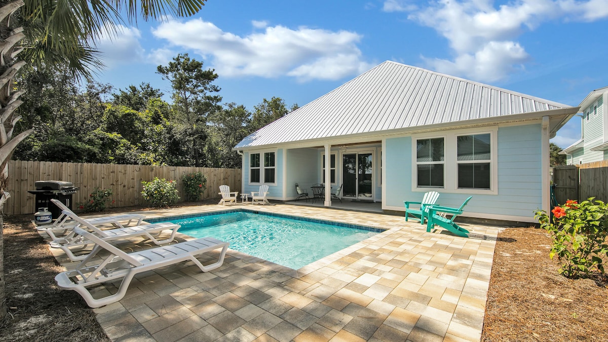A private pool is surrounded by a spacious patio, featuring lounge chairs and colorful outdoor seating. The house, with a light blue exterior and a metal roof, overlooks the pool. Lush greenery and a wooden privacy fence frame the area, creating a tranquil outdoor setting.