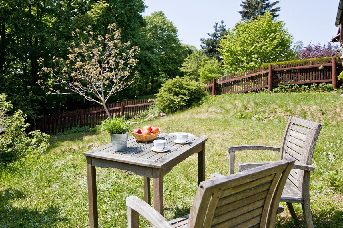 An outdoor seating area is presented with a wooden table and two chairs, inviting relaxation. A bowl of apples sits on the table, surrounded by lush greenery and a flowering tree. A rustic fence encloses the garden, enhancing the private atmosphere.