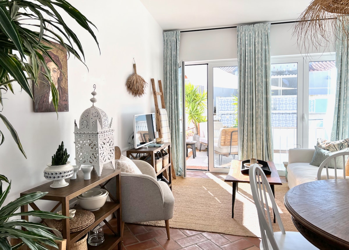 A bright living space features a light-colored sofa and stylish decor. Large sliding doors lead to a sunny terrace, enhancing the open atmosphere. Textured curtains frame the windows, while potted plants and decorative items add warmth and character to the room.