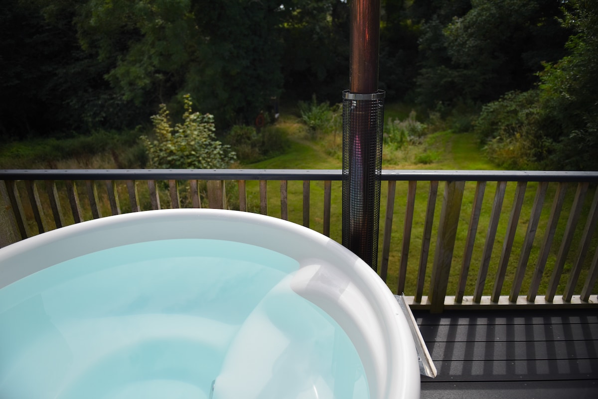 A private wood burning hot tub is seen on a wooden deck, surrounded by lush greenery. The tranquil setting overlooks a garden area, with trees and shrubs providing a natural backdrop. A heater stands nearby, ensuring comfort during cooler evenings.