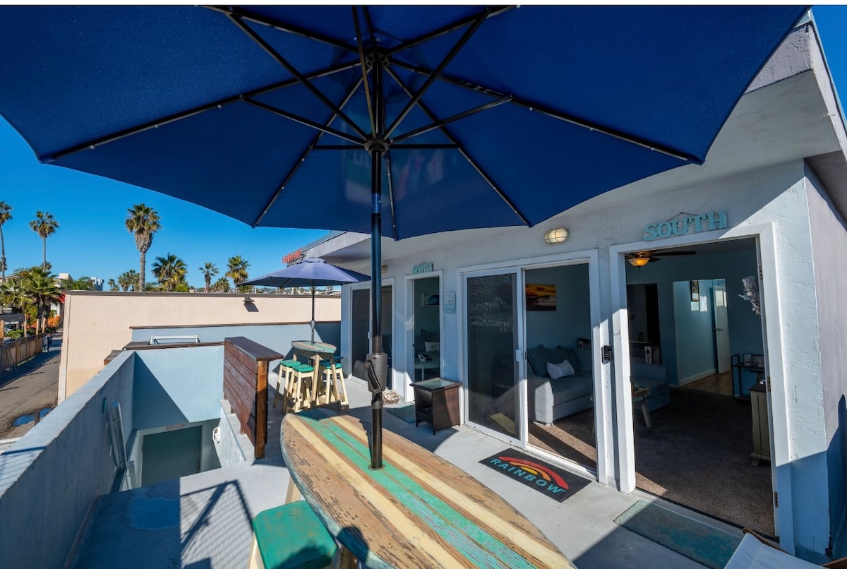 The private rooftop deck features a surfboard table beneath a large blue umbrella. Comfortable seating options are arranged for outdoor relaxation. Views of palm trees and nearby structures are visible, emphasizing a relaxed coastal atmosphere.