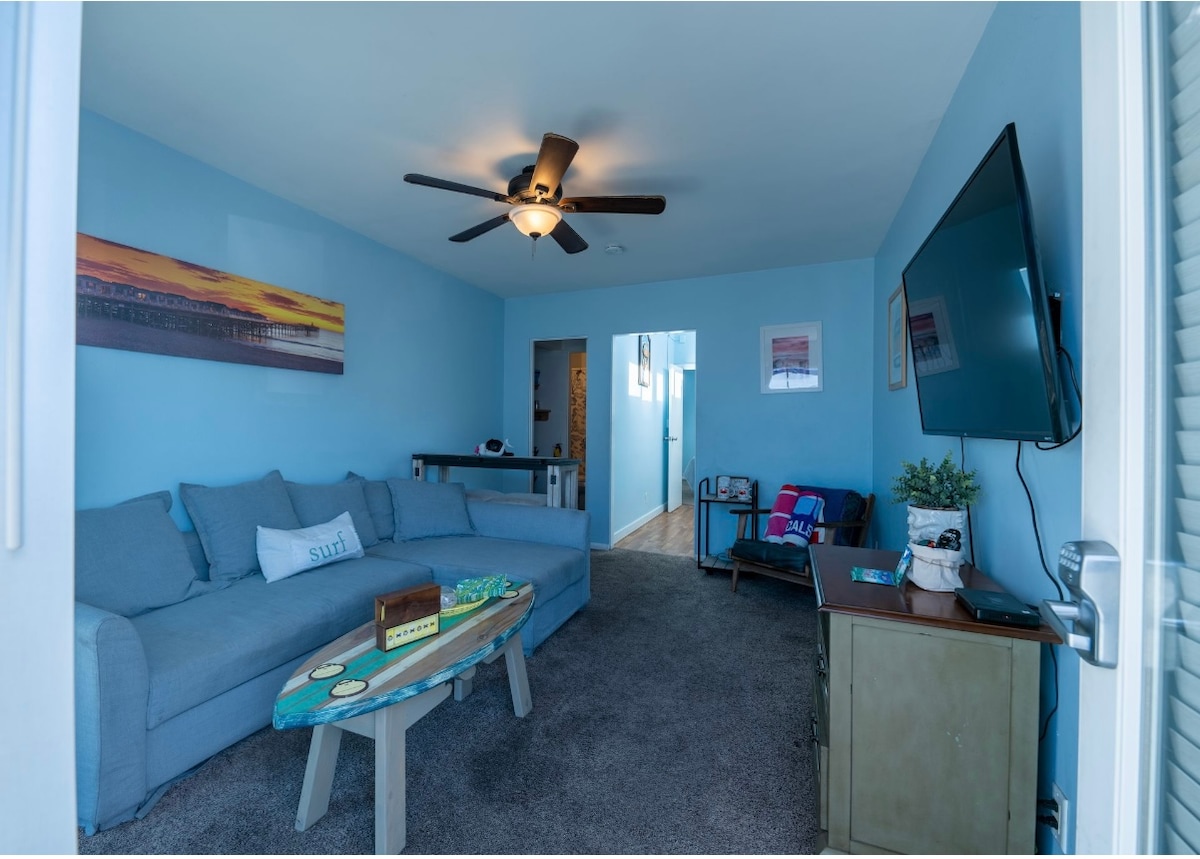 A light blue living room features a spacious couch and a surfboard coffee table, enhancing the beach-themed decor. A ceiling fan is mounted for air circulation, and a wall-mounted television is visible. Doorways lead to other areas, and a small table is positioned against the wall.