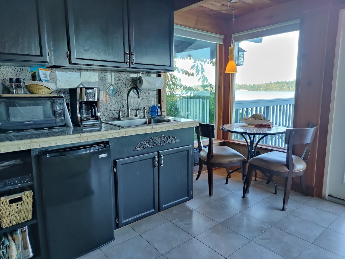 The kitchen area features dark cabinetry complemented by a speckled countertop. A small table with two chairs is positioned near a window, offering a view of the water. Appliances include a microwave and coffee maker, providing essential amenities for convenience.