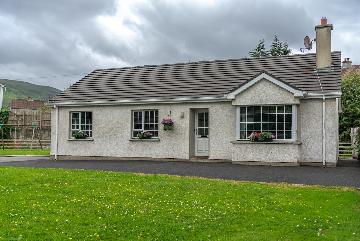 A single-story detached holiday home is shown, featuring a light-colored exterior with multiple flower boxes beneath large windows. A well-maintained lawn leads to the front door, which is white with a simple design. The sky displays varying shades of gray.