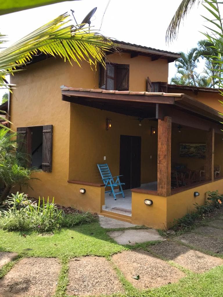 A two-story chalé features a warm yellow exterior with wooden accents. The entrance is framed by lush greenery and a pathway made of stone tiles. The porch is furnished with a blue chair and illuminated by soft outdoor lighting.