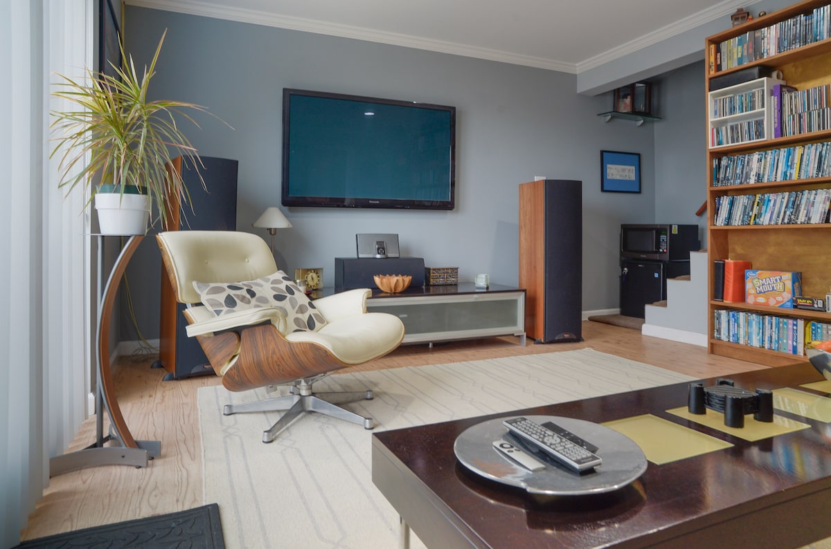 A spacious living area features a comfortable lounge chair alongside a sleek coffee table. A large flat-screen TV is mounted on the wall, while tall speakers flank the room. A wall unit filled with DVDs adds a cozy touch, complemented by natural light from large windows.
