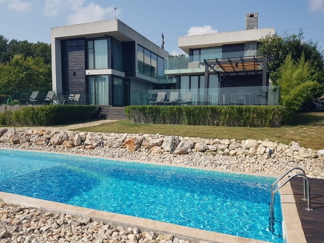 A modern house is showcased with large glass windows reflecting natural light. A clear swimming pool runs parallel to the house, surrounded by a landscaped garden featuring stone paths and greenery. Sun loungers are positioned on a spacious terrace, ready for relaxation.