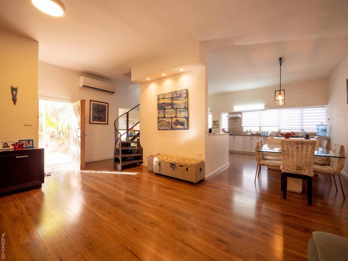 A spacious living area features warm wooden flooring, with an inviting dining table surrounded by woven chairs. Natural light enters through large windows, illuminating a nearby kitchen area. An artistic wall piece adds a touch of character to the open layout, promoting a welcoming atmosphere.