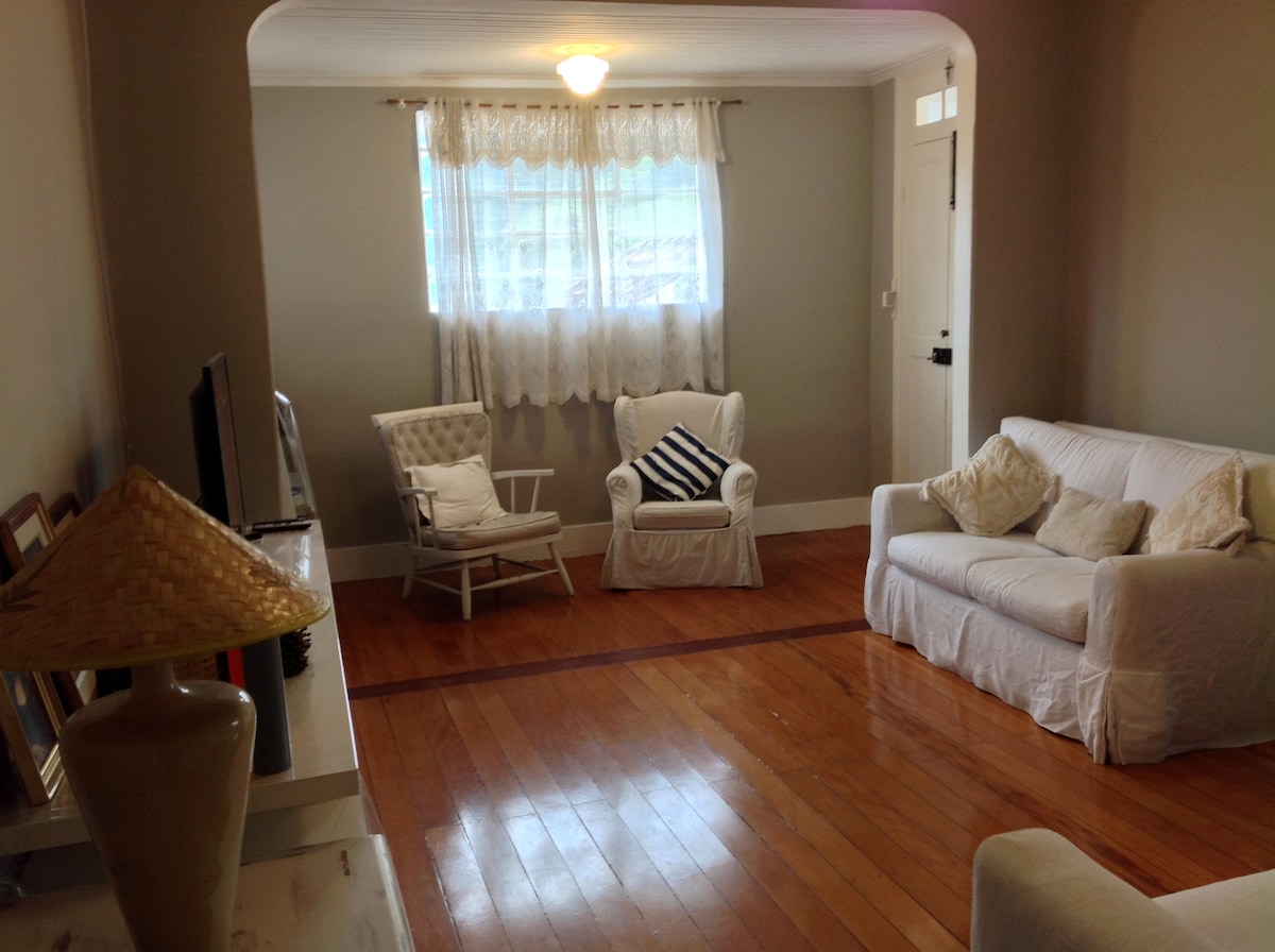 A bright and spacious living area showcases polished wooden floors and natural light filtering through a window with sheer curtains. The space is furnished with white sofas, a decorative lamp, and a comfortable armchair, creating a welcoming environment for relaxation.