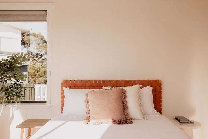 Cozy bedroom featuring a queen bed with a woven headboard and soft, textured pillows. Large window brings in natural light, creating a peaceful and relaxing atmosphere.