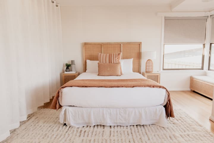 Spacious bedroom featuring a queen bed with a woven headboard, soft linens, and warm-toned pillows. The neutral decor, large windows, and cozy rug create a calm and inviting atmosphere.