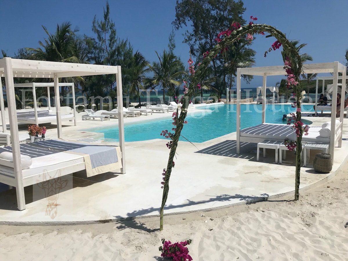 An inviting pool area features white lounge beds framed by tropical palm trees, creating a peaceful atmosphere. A decorative arch adorned with vibrant flowers leads the eye towards the shimmering blue water, enhancing the serene environment around the expansive pool.