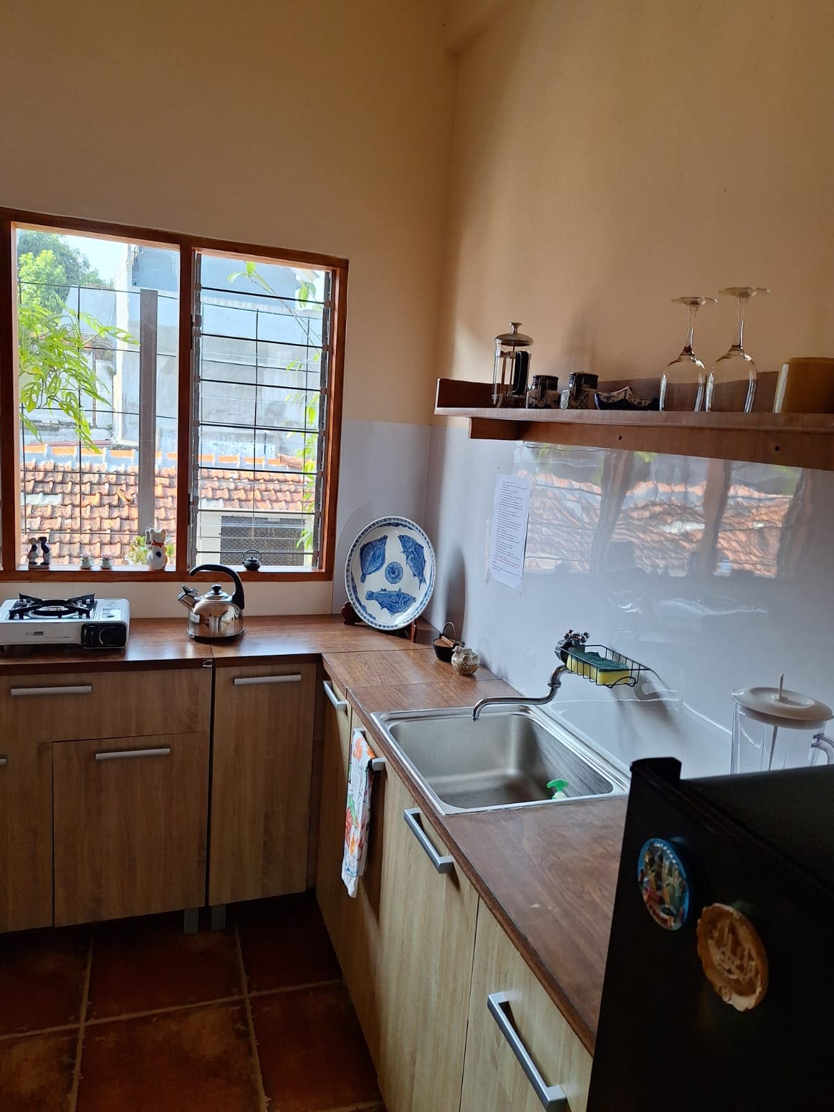 A well-equipped kitchen is shown, featuring wooden cabinetry and a stainless steel sink. A gas stove sits beside a refrigerator. Natural light enters through a window, providing a glimpse of greenery and tiled roofs outside. Decorative items are arranged on shelves.