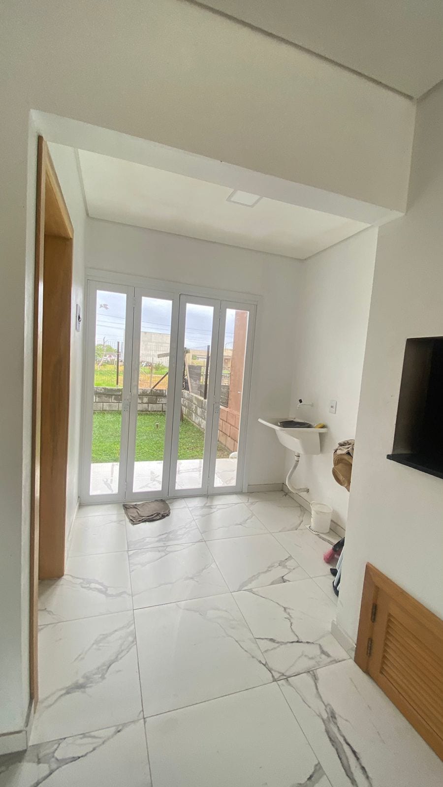 A bright entryway features large glass doors that provide access to a green outdoor space. Light marble tiles cover the floor, and a small sink is mounted on the wall, enhancing the practical layout of the room.