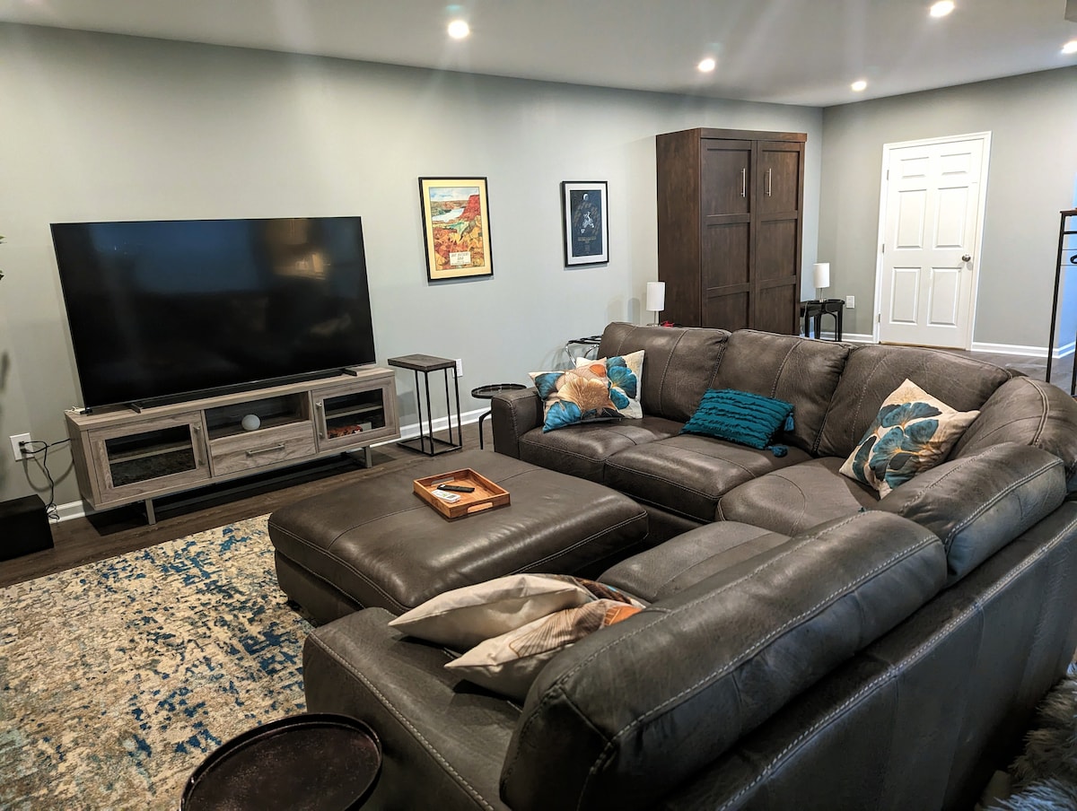 A spacious living area features a large sectional sofa adorned with decorative pillows. A large television is mounted on a media console, and artwork is displayed on the walls. The room is illuminated by overhead lighting, providing a welcoming environment for relaxation.