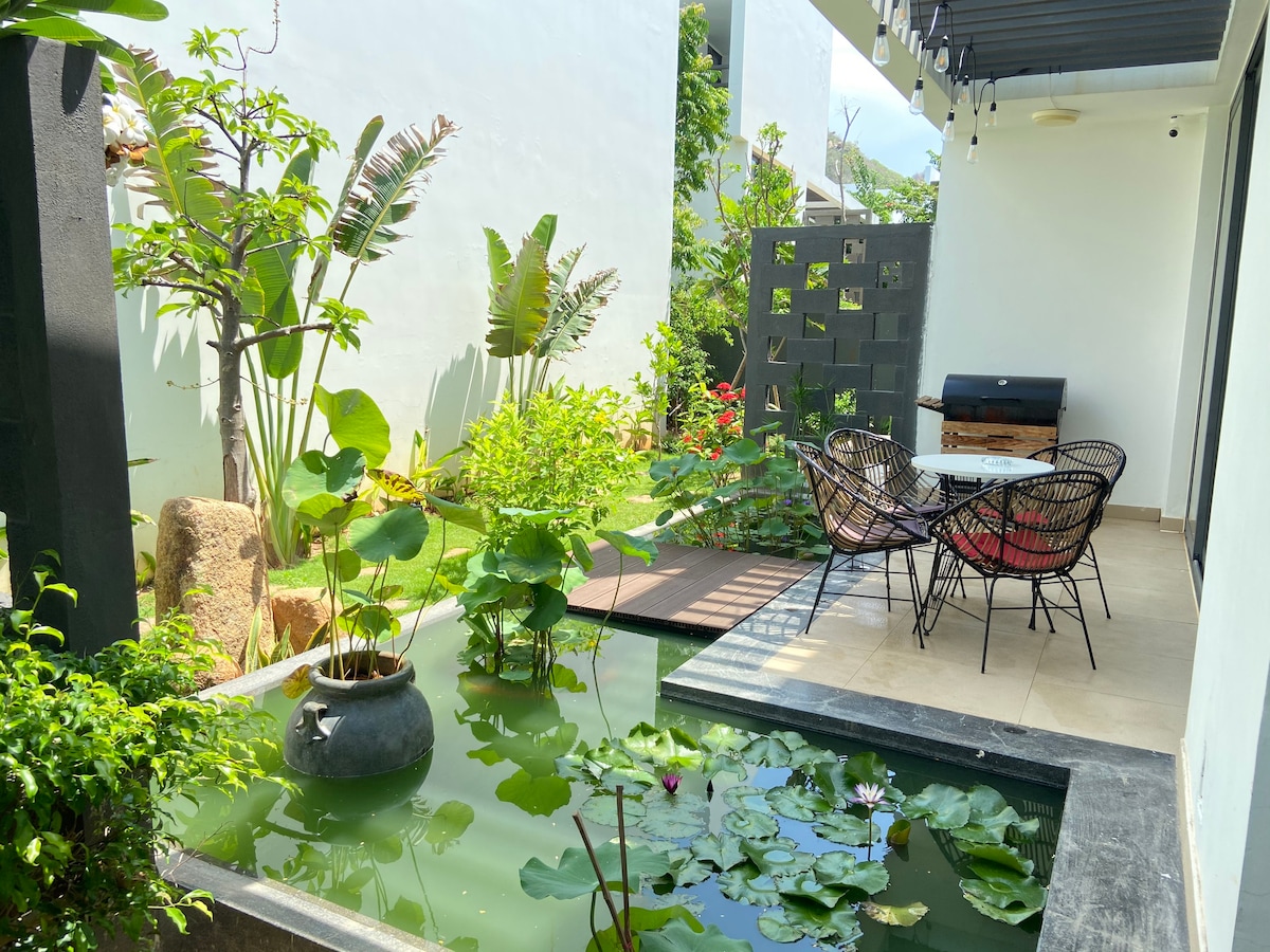 A tranquil outdoor space includes a serene pond with floating lily pads and a decorative pot. A table with four chairs is positioned on a tiled patio, surrounded by lush greenery and colorful plants. Subtle sunlight filters through the overhead pergola.