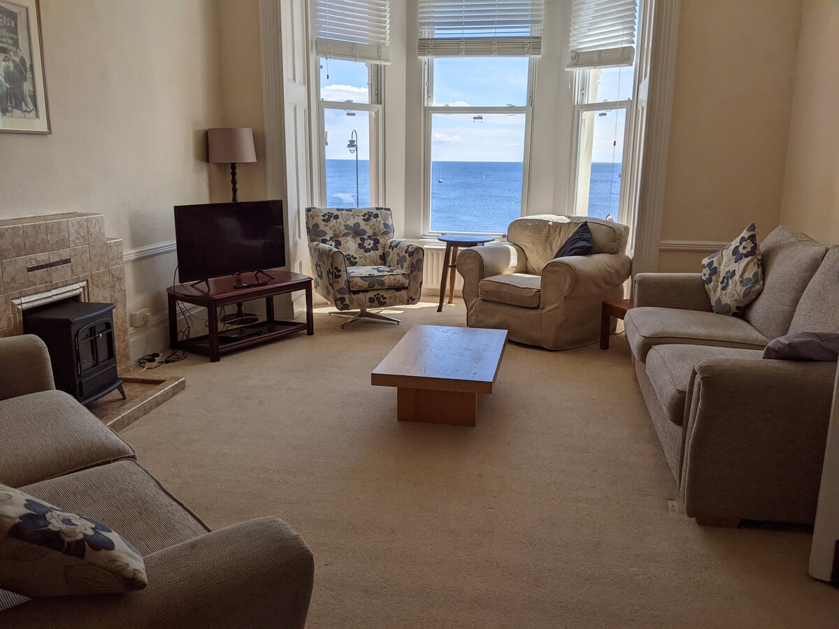A shared lounge area is shown, featuring comfortable seating arrangements including a beige sofa and patterned armchairs. A wooden coffee table occupies the center, while a television is positioned against the wall. Large windows invite natural light and offer views of the seaside.