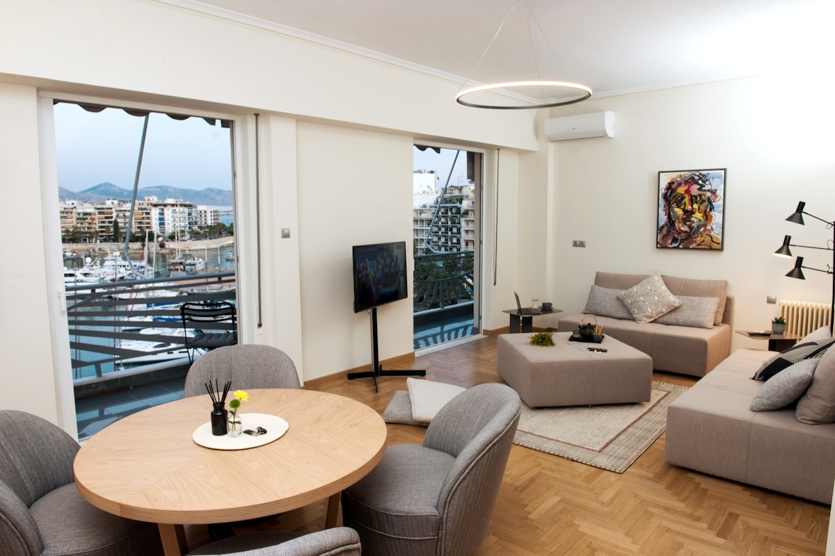 A spacious living-dining area features a circular wooden table surrounded by four gray chairs. Two comfortable sofas are positioned near large windows, offering views of the marina. A television is mounted on the wall, and tasteful artwork decorates the space, enhancing the modern aesthetic.