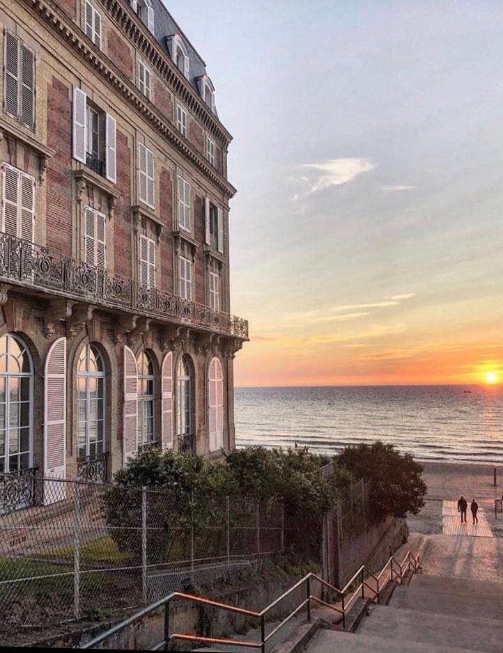 The image captures the historic exterior of the building at sunset, featuring large arched windows and ornate metal railings. A landscaped area is visible in the foreground, and the beach is directly accessible, with waves gently lapping at the shore in the background.