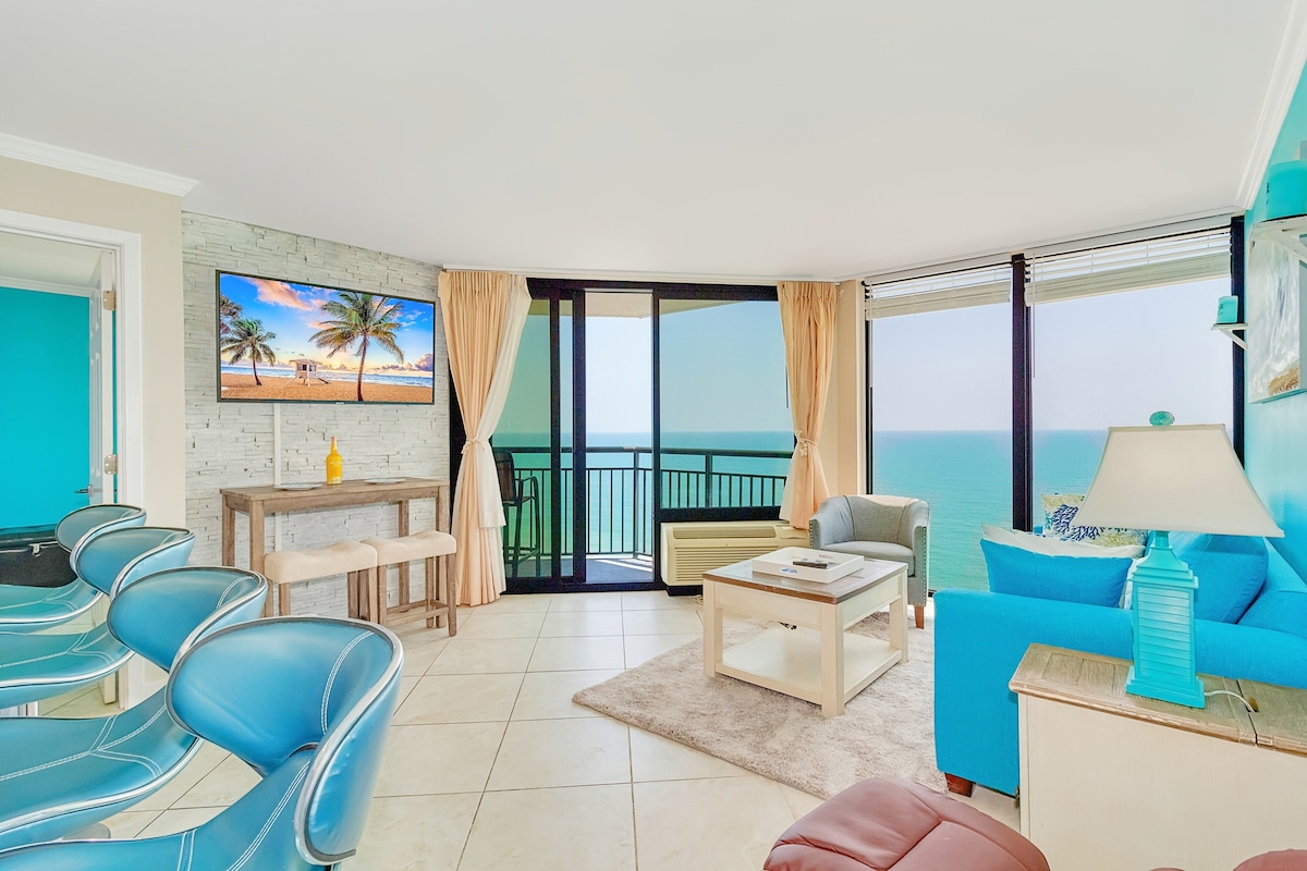 The living area features floor-to-ceiling glass windows providing an unobstructed view of the ocean. A cozy seating arrangement includes a blue sofa and two contemporary chairs. A flat-screen TV is mounted on a textured stone wall, creating a bright and airy ambiance.