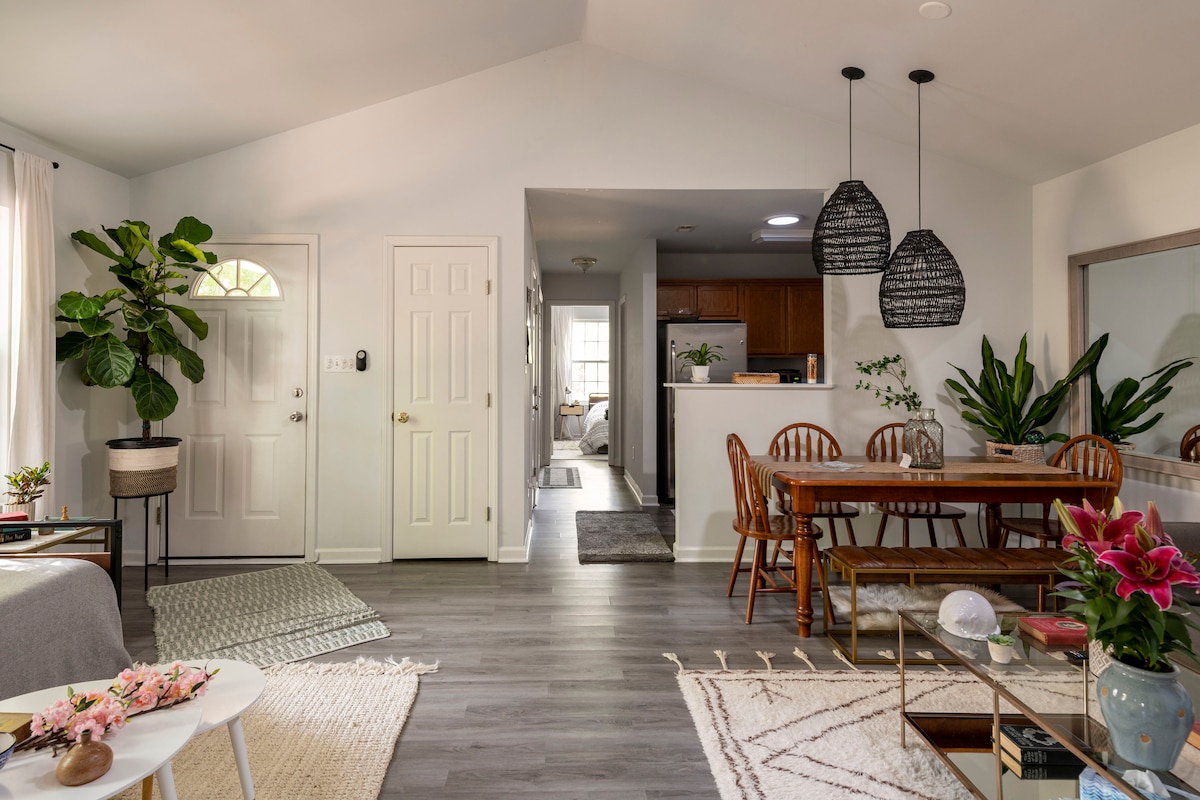 An open living and dining area features a cozy seating space with a light grey sofa, a decorative plant, and a large dining table with wooden chairs. Natural light floods in through large windows, while a modern kitchen is visible in the background.