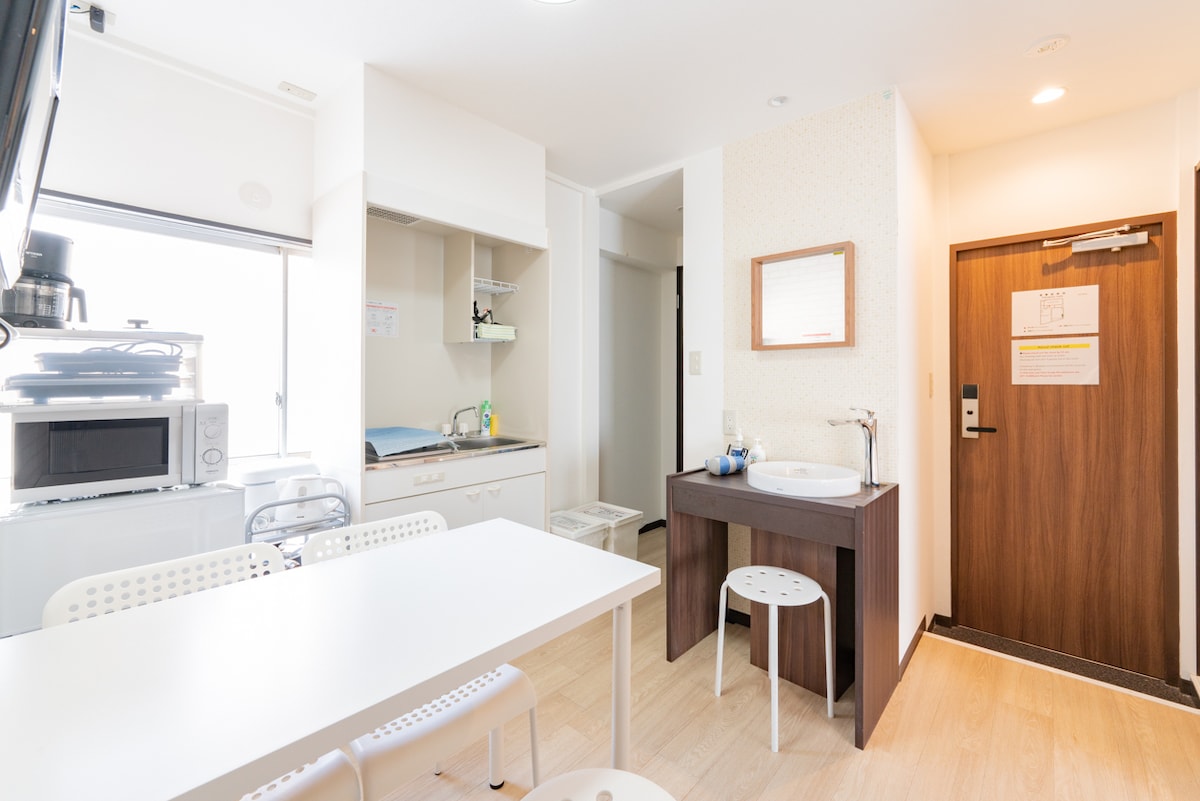 A bright and functional kitchen area is presented, featuring a white dining table with four chairs. A small washbasin and mirror are positioned nearby, with a microwave and other appliances visible in the background. Natural light enters through a window, enhancing the space's warmth.