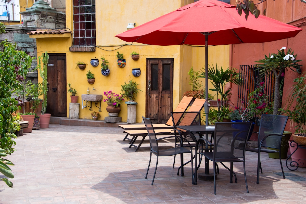 A shared terrace features a large red umbrella providing shade over a dining table surrounded by several chairs. Potted plants add greenery to the space, and the walls display colorful flower pots. Two loungers are positioned nearby, creating a relaxing outdoor area.