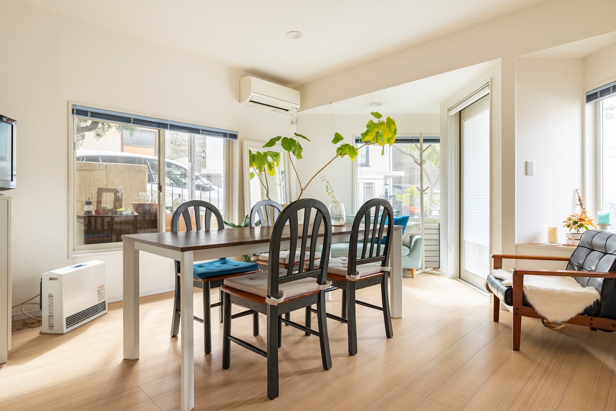 A spacious dining and living area is visible, featuring a wooden dining table with six black chairs. Large windows provide ample natural light, and a cozy seating arrangement is located in the corner. Green plants add a touch of nature to the space.