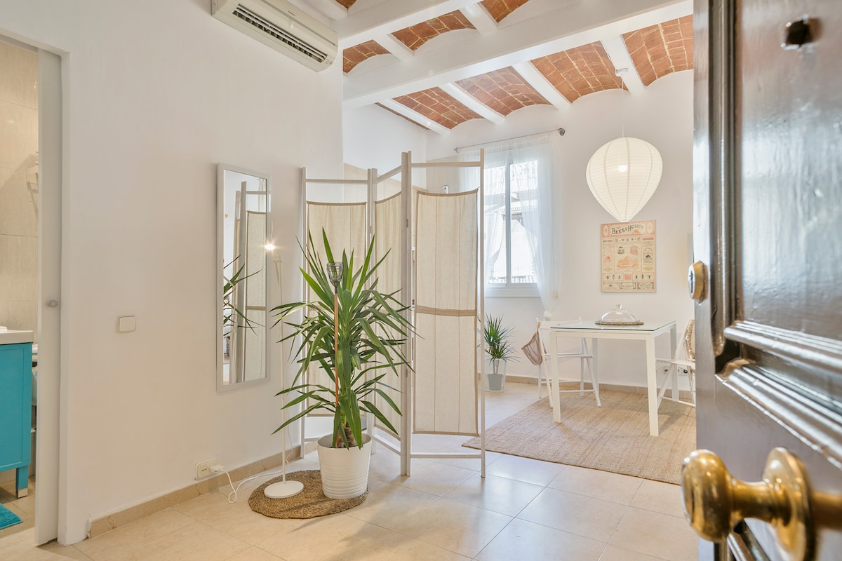 A welcoming entryway is showcased, featuring a decorative folding screen and a tall green plant in a white pot. Bright light filters through a nearby window, illuminating a small dining table and chair arrangement. Exposed ceiling beams enhance the tranquil atmosphere.