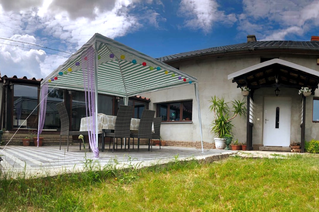 An outdoor pavilion is seen, featuring a dining set with several chairs and a table. Colorful lights are draped along the top, providing a festive touch. The surrounding garden is well-kept, with a mix of grass and potted plants, completing a welcoming outdoor space.