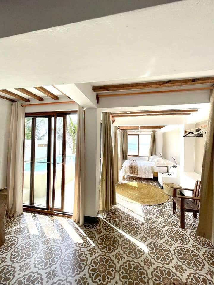 A bright interior showcases an inviting space with a view of the private plunge pool. Natural light filters through large glass doors, illuminating the patterned tile floor and modern furnishings. A cozy sleeping area is visible, featuring a bed dressed in crisp white linens.