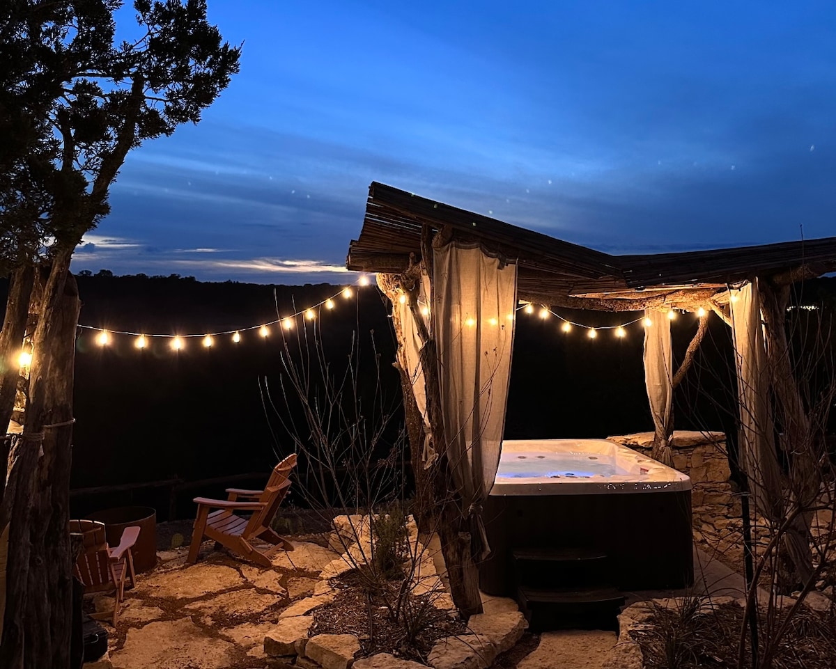 The outdoor hot tub is surrounded by soft lighting, enhancing the serene evening ambiance. A natural stone patio offers a rustic feel, complemented by wooden chairs positioned nearby. The sky features subtle gradients of blue, indicating dusk in a tranquil setting.