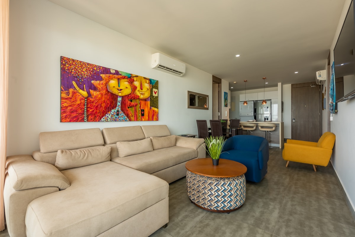 A spacious living area features a light-colored sectional sofa and a round wooden coffee table. Bright artwork adorns the wall, contributing color to the space. An inviting armchair in a vibrant yellow is positioned nearby, while the open kitchen and dining area are visible in the background.