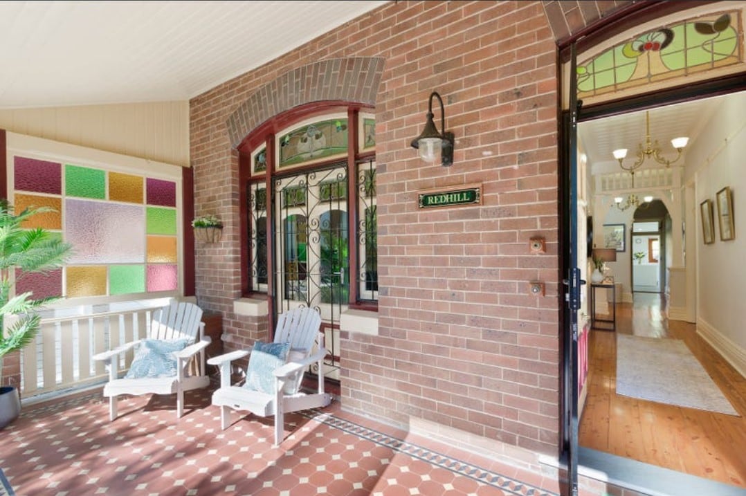 A welcoming front porch features two white adirondack chairs positioned on a patterned tiled floor. Surrounding the entrance, stained glass windows with colorful panels create a cheerful atmosphere. The doorway leads to an inviting interior with natural light flowing through.