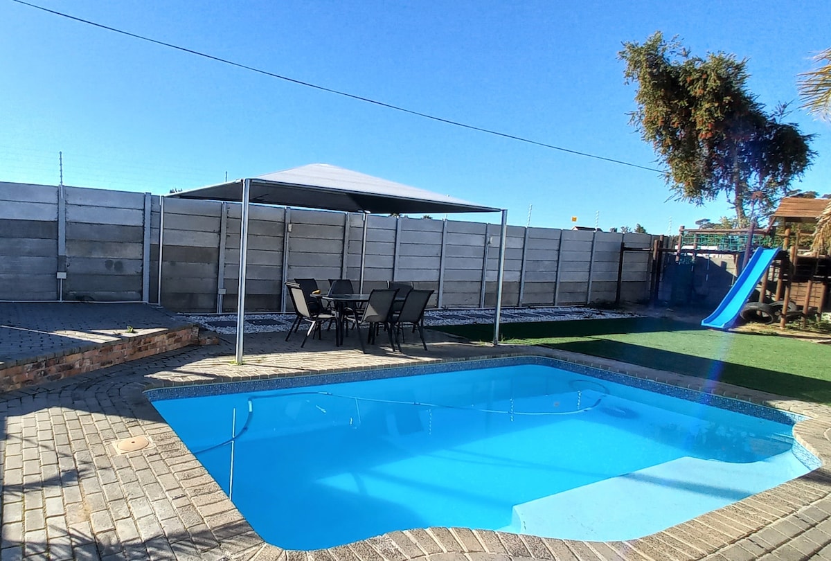 A private pool is surrounded by a paved area featuring a shaded seating arrangement of six metal chairs and a table. Adjacent, a grassy area leads to a blue slide. The backdrop consists of a secure fence under a clear blue sky.