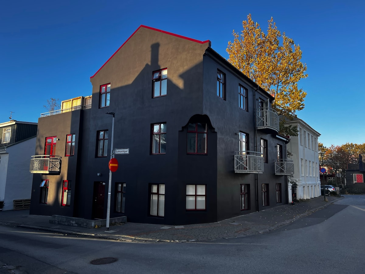 A modern three-story building with a dark exterior, highlighted by red window frames and balconies. The structure stands at the intersection of two quiet streets, with trees shedding autumn leaves in the foreground, creating a peaceful urban setting.