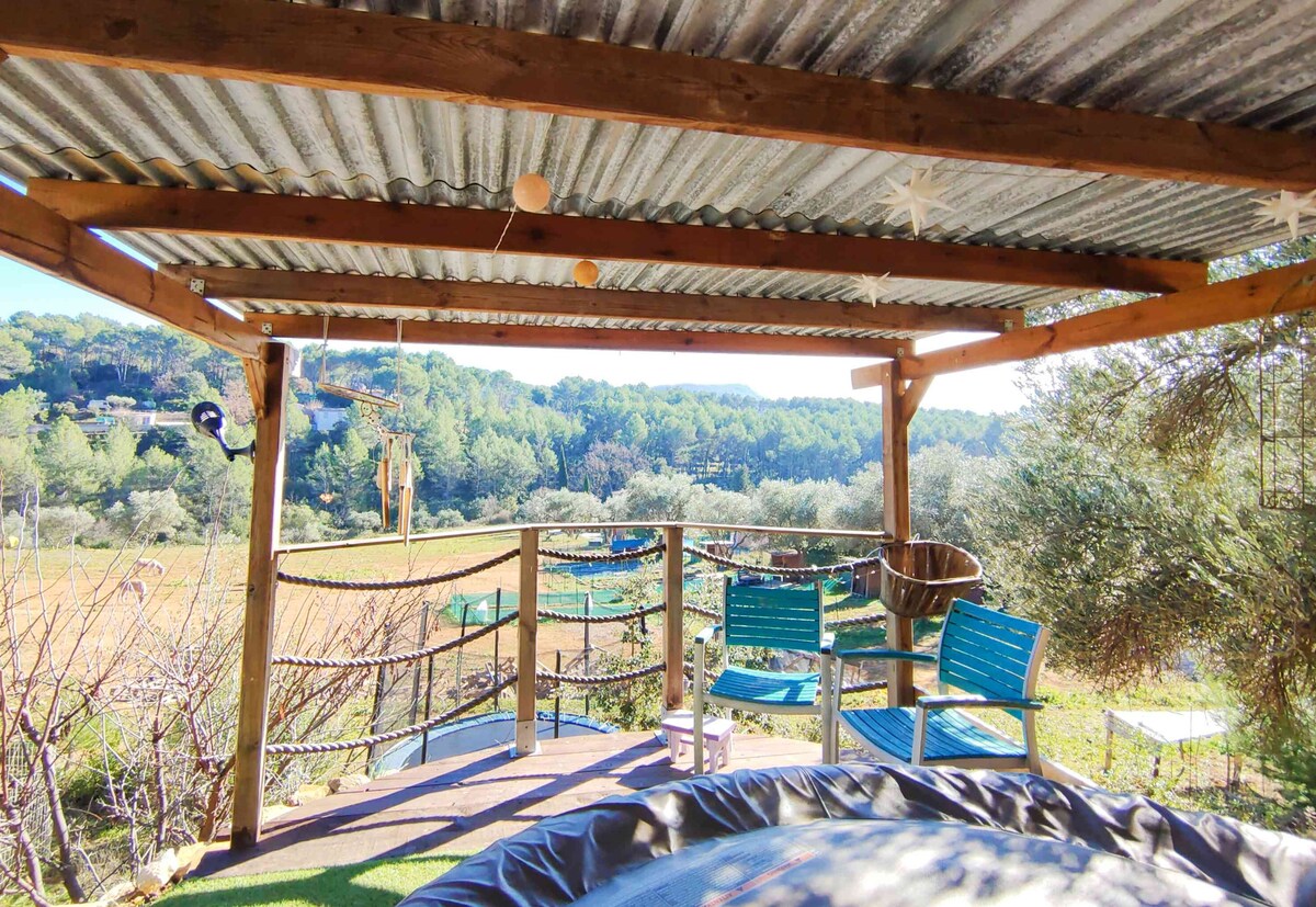 A wooden terrace is covered by a corrugated roof, offering a shaded area with views of the surrounding greenery. Two teal chairs are positioned on the deck, and a trampoline is visible below, surrounded by a natural landscape.