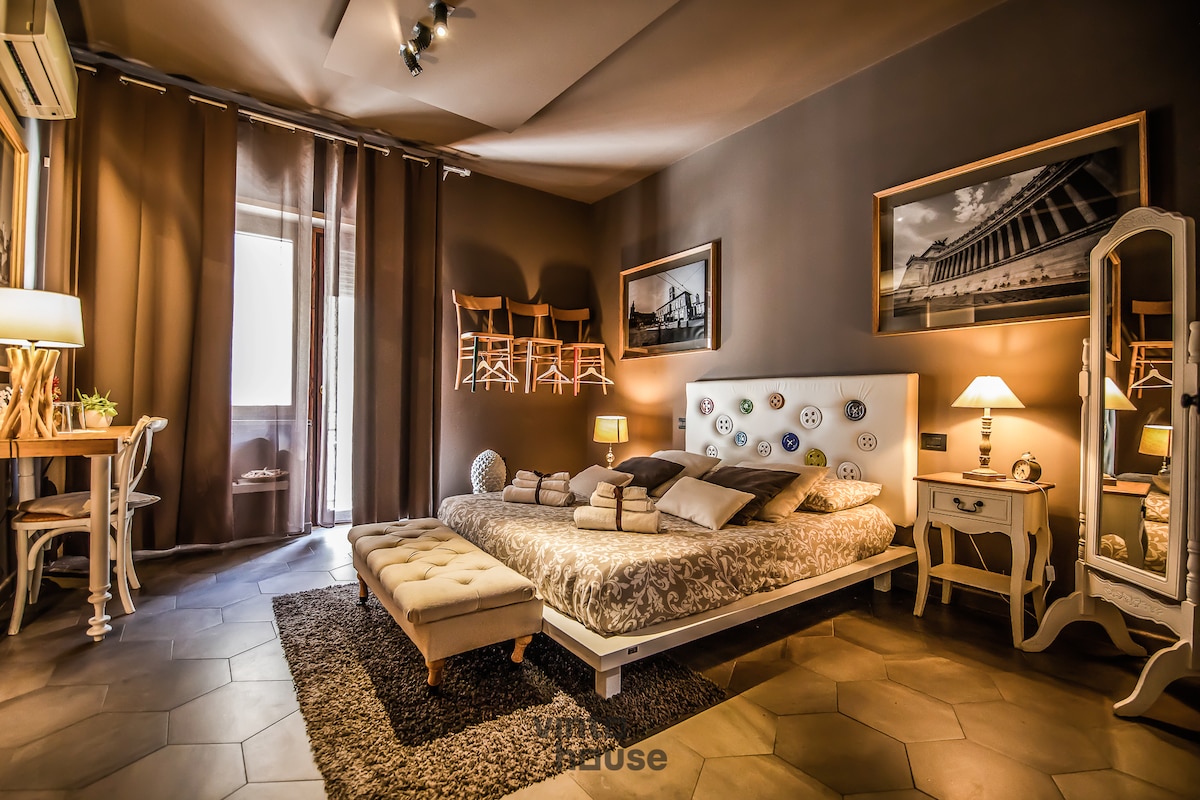 A spacious bedroom is arranged with a large bed, adorned with soft pillows and a decorative throw. Natural light filters through sheer curtains, creating a warm ambiance. Decor includes a vintage mirror and artwork on the walls, complemented by a cozy seating area and a small dining table.