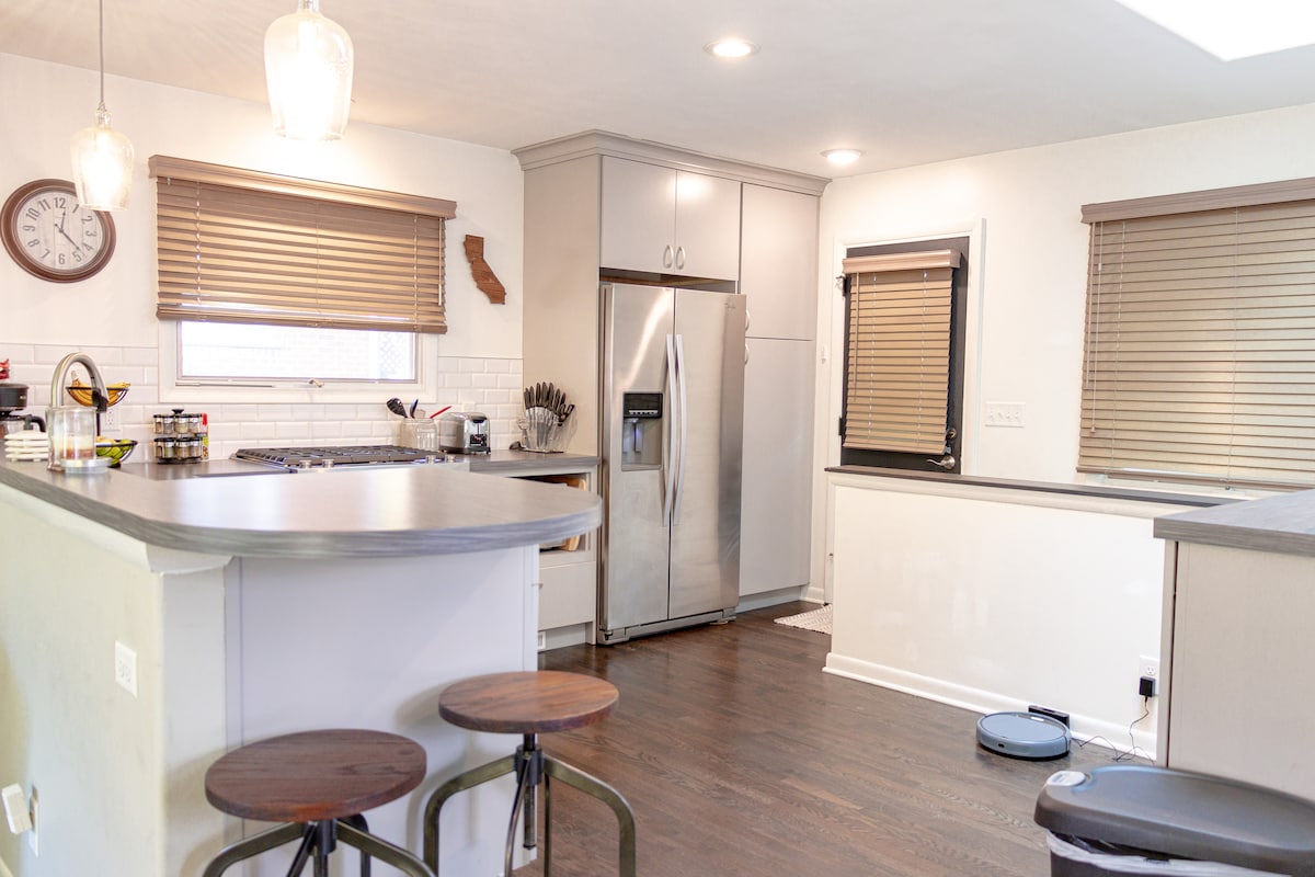 The kitchen features modern appliances, including a stainless steel refrigerator and a gas stove. Counter space is provided along with two wooden bar stools at the island. Blinds cover the windows, allowing natural light to fill the room.