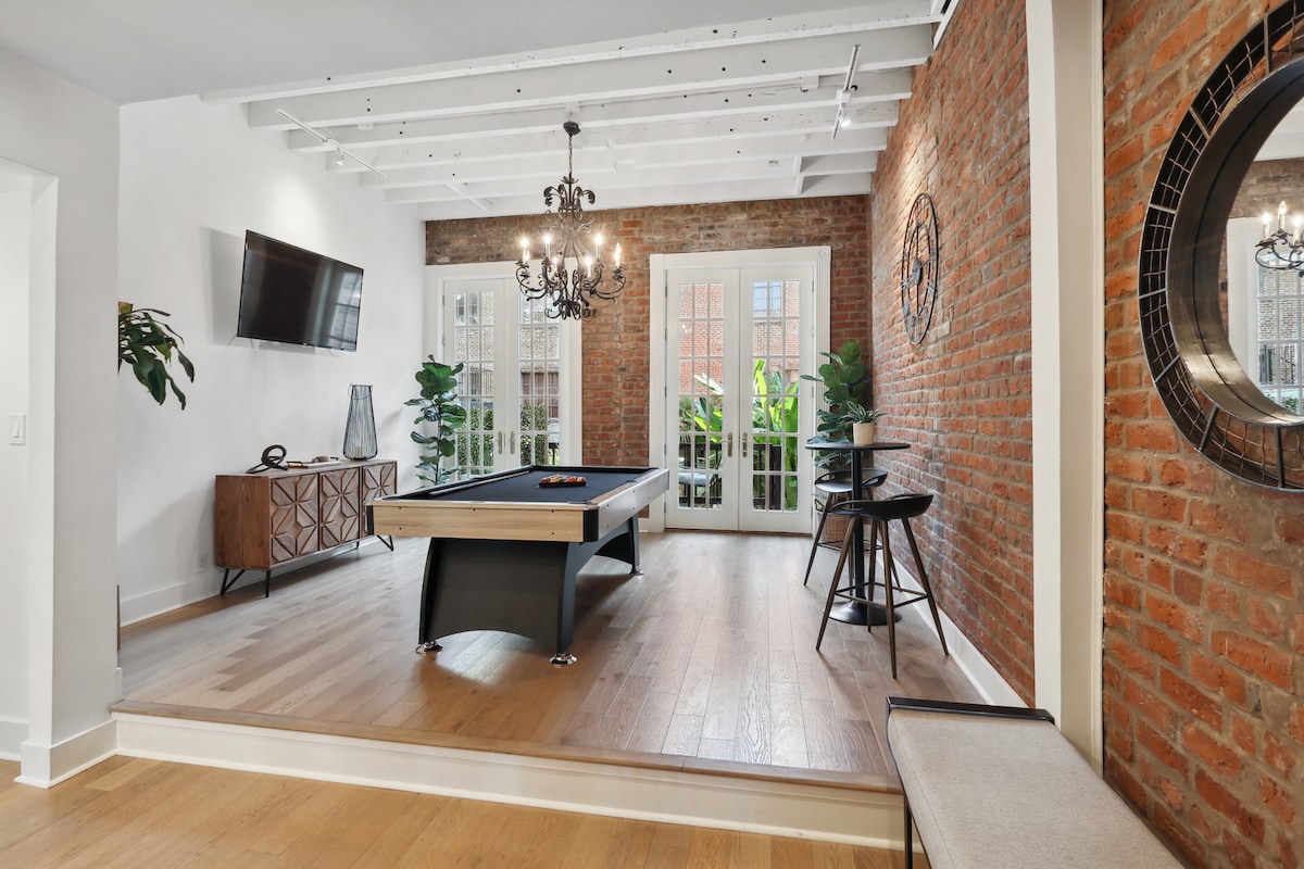 A pool table is positioned within a stylish room featuring exposed brick walls and wooden beams. Large glass doors lead to a balcony, enhancing natural light flow. Potted plants are visible, contributing to the room's vibrant ambiance, along with a modern chandelier and wall-mounted TV.