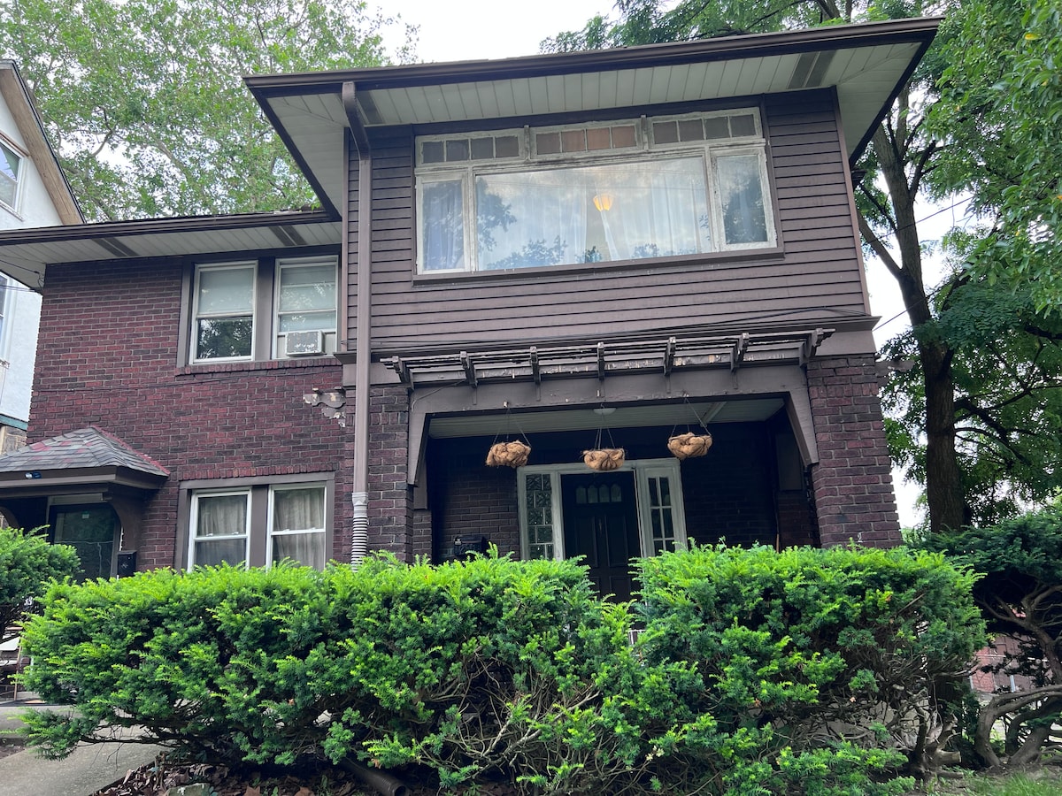 A charming two-story brick building with a distinctive overhang above the entrance. Large windows allow natural light to illuminate the interior, while a green hedge frames the front yard, providing a sense of privacy. The entrance is adorned with hanging planters.