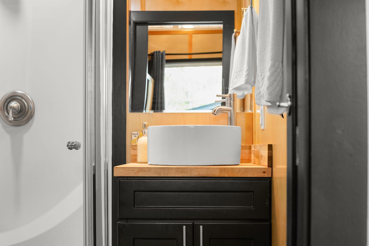 The bathroom features a modern design with a wall-mounted sink atop a wooden surface. A mirror is above the sink, and two towels are neatly hung nearby. The shower is visible in the background, showcasing a clean and functional space.