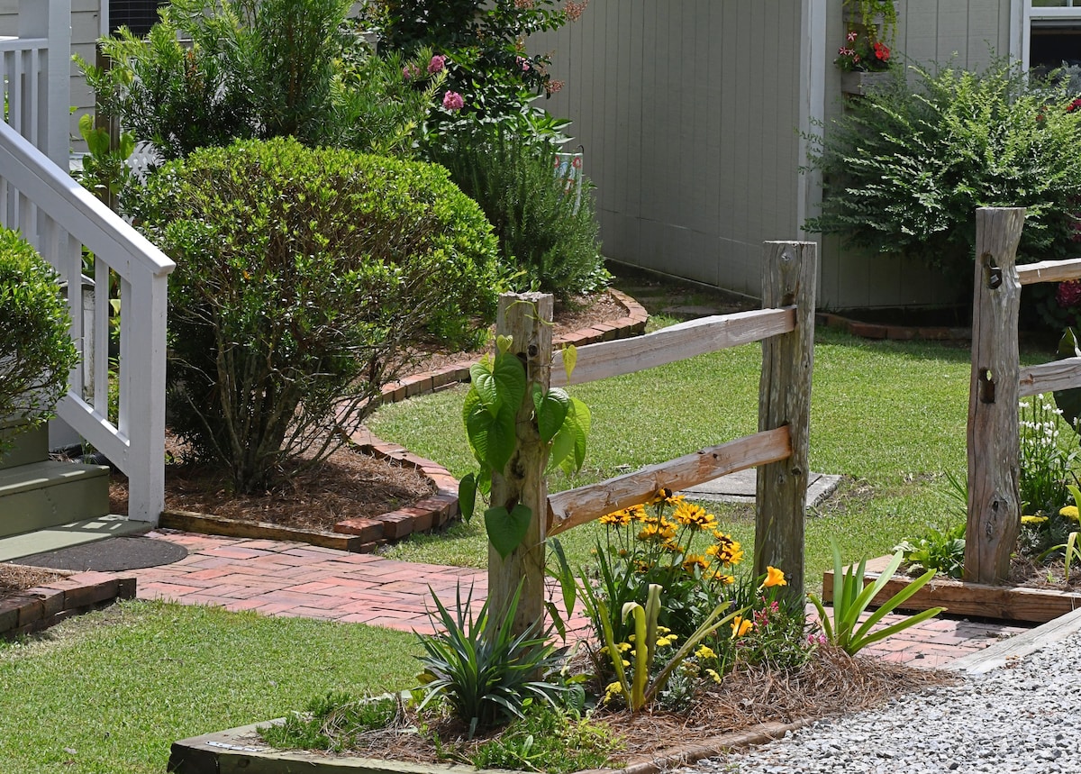 A well-maintained garden area features an assortment of vibrant flowers and lush green shrubs. A sturdy wooden fence stands beside a landscaped pathway, inviting guests to explore the serene outdoor space. The manicured lawn is complemented by a nearby gravel driveway, enhancing the natural beauty.