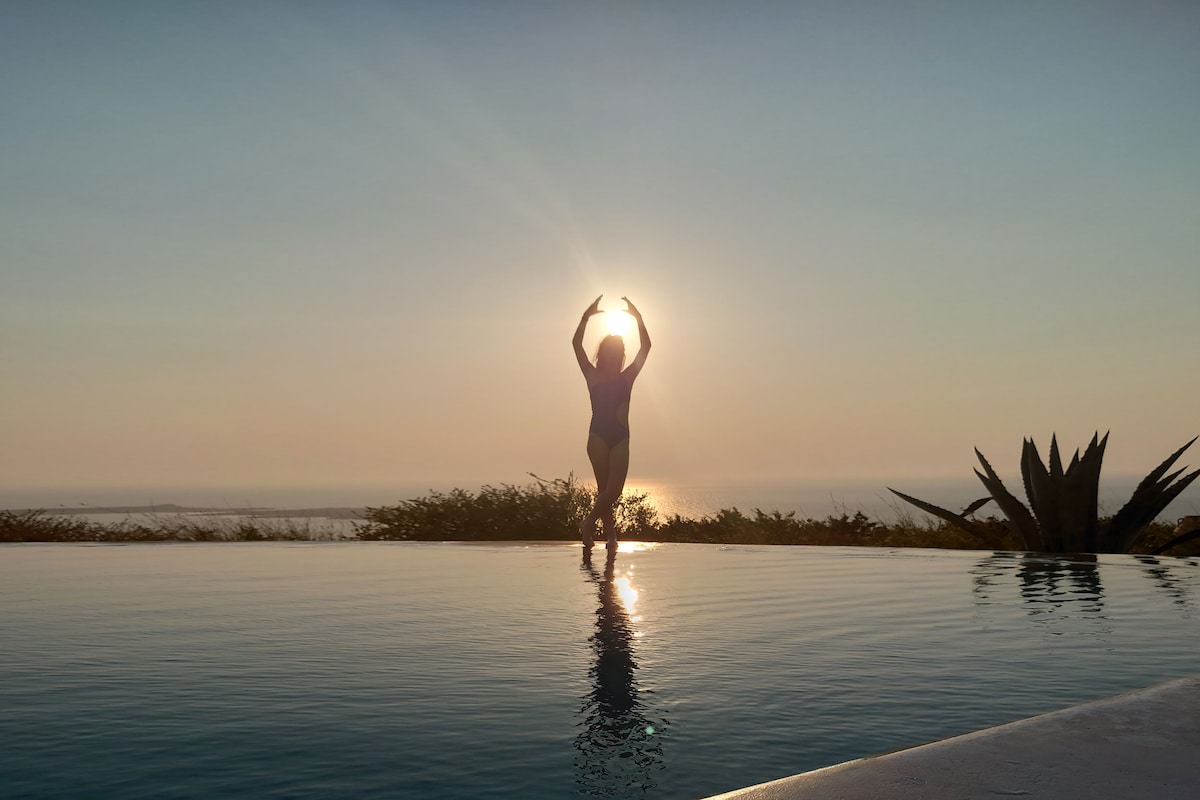 An individual is silhouetted against the rising sun while standing in an infinity pool, with a view of the coastline and distant islands. The scene captures tranquil water reflecting the sky hues and lush vegetation surrounding the pool area.