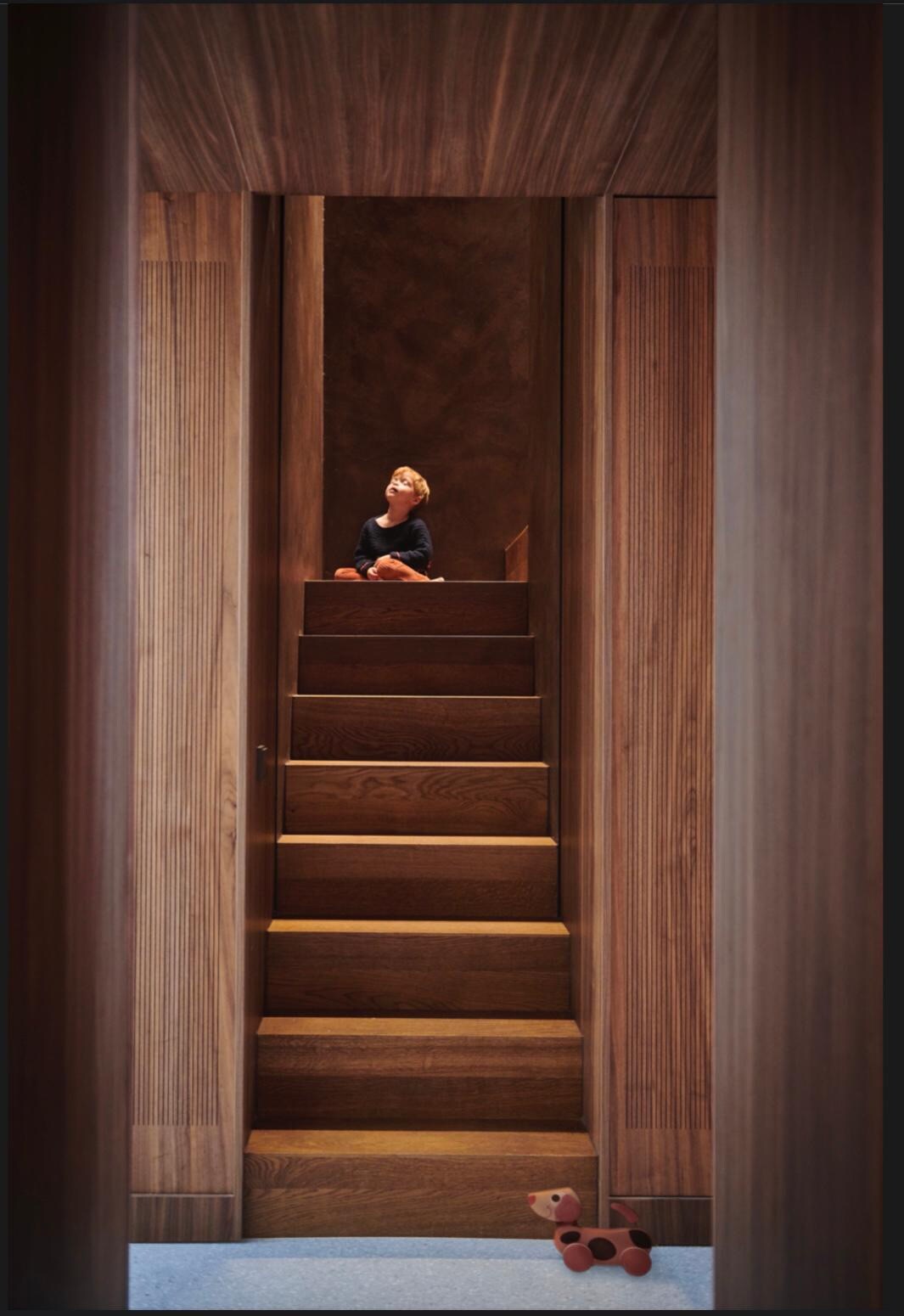 A staircase made of warm wooden steps leads upward, framed by wooden walls that create a cozy tunnel effect. A small toy is positioned on the floor at the bottom of the stairs, and a figure is seen sitting at the top.