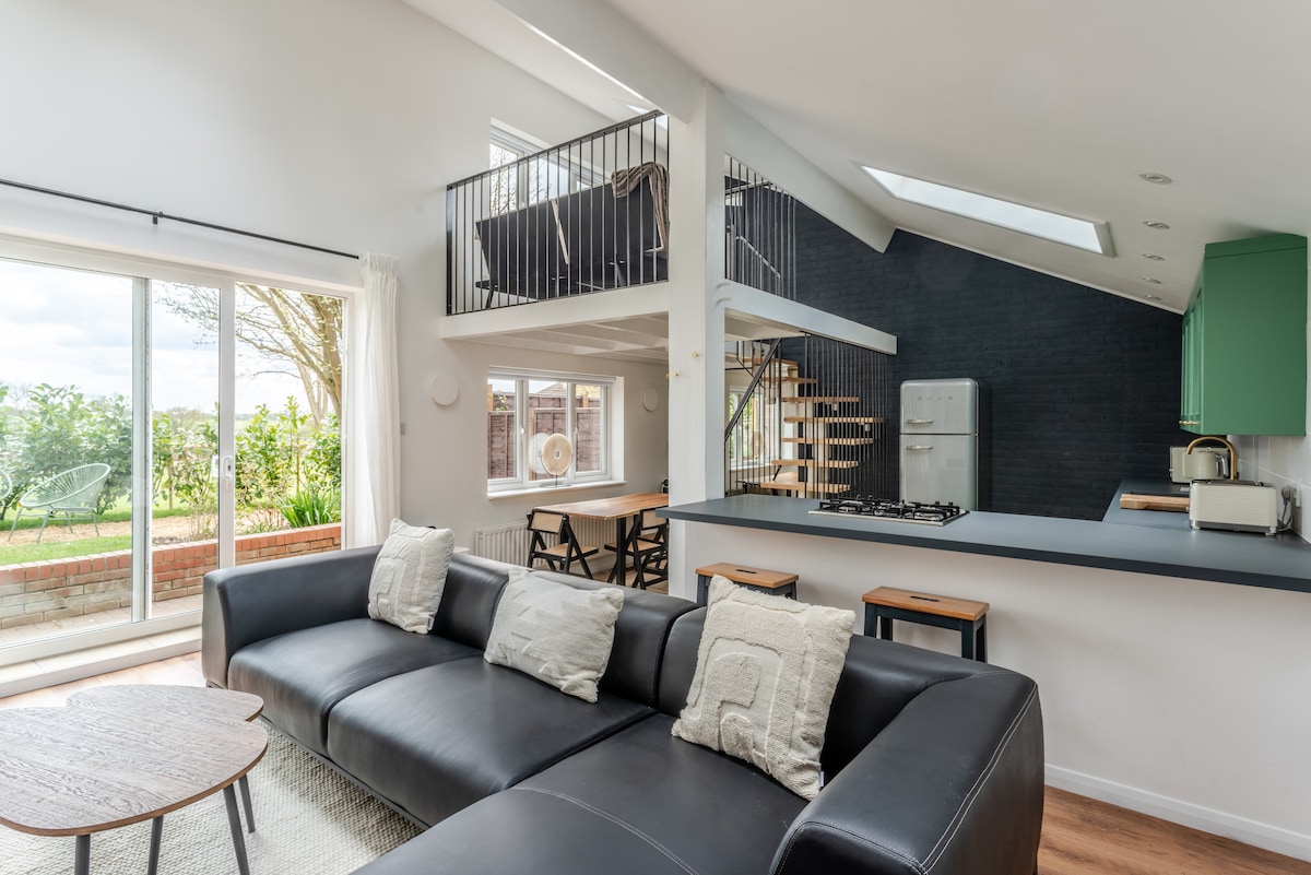 A spacious living area features a modern black sectional sofa, complemented by a coffee table. Large glass doors lead to the garden, while a staircase ascends to the mezzanine level. The kitchen area, highlighted in green, is equipped with contemporary appliances.