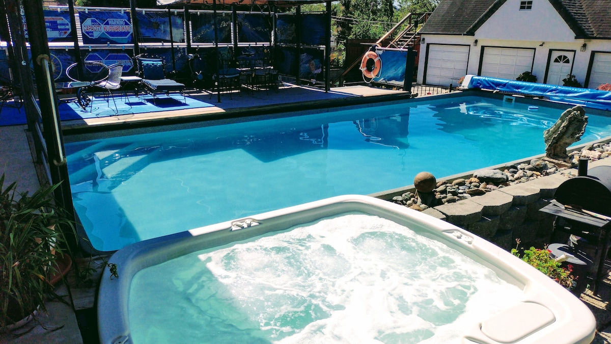 A spacious pool area is visible, featuring a large, clear blue swimming pool alongside a bubbling hot tub. Surrounding areas include lounge chairs and a deck with an unobstructed view of the water. Lush landscaping and stone details add a natural touch.