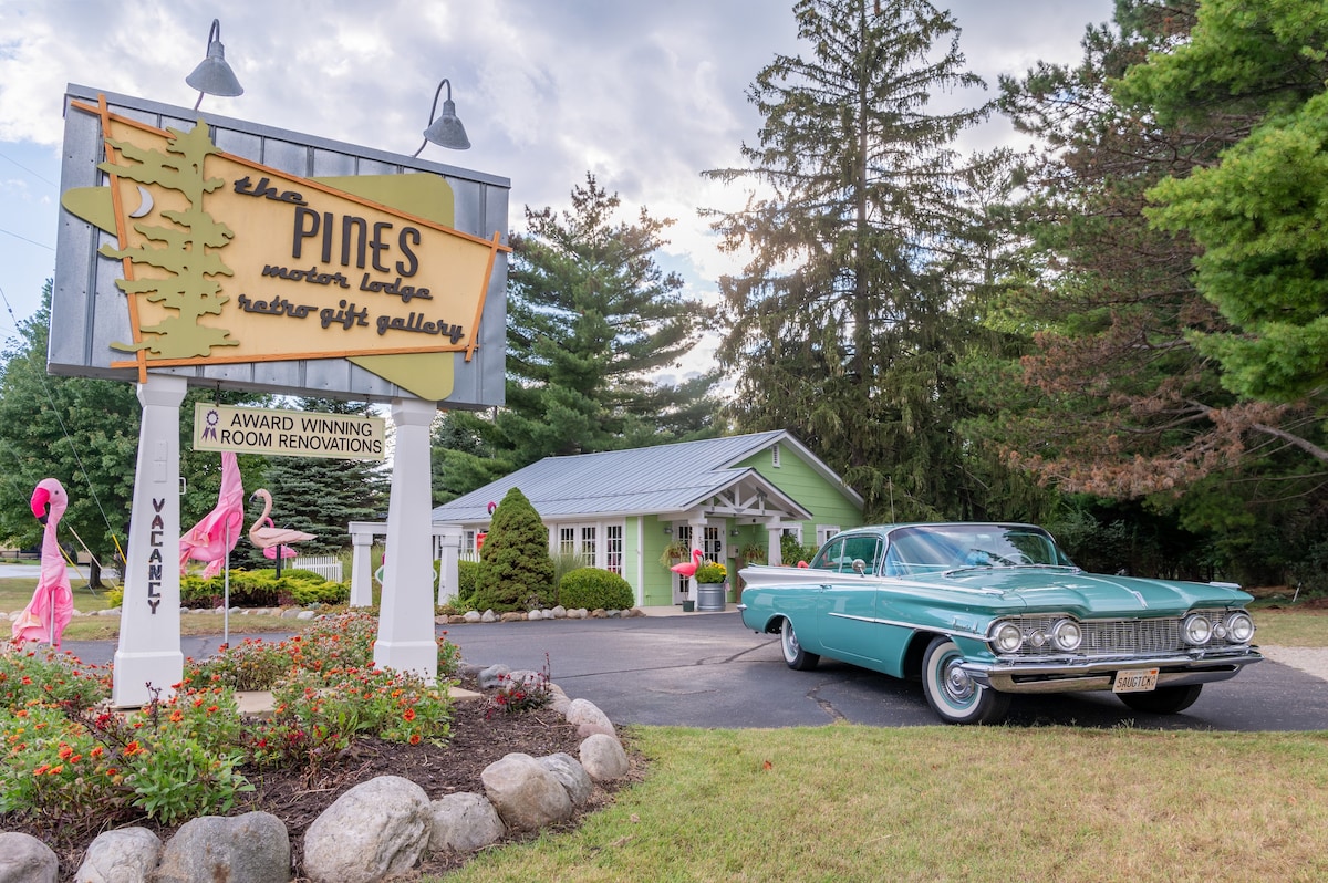 The welcoming entrance of the Pines is showcased, featuring a vibrant sign announcing its award-winning renovations. A classic turquoise car is parked in front, surrounded by landscaped greenery and boulders, contributing to a charming and accessible atmosphere.