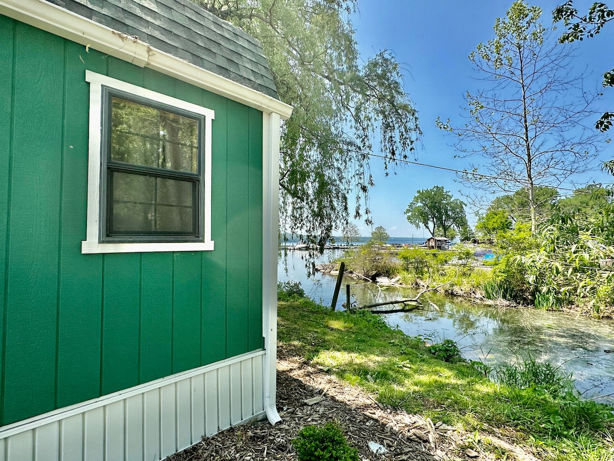 The cabin's vibrant green exterior is framed by a white trim, with a large window reflecting the surrounding nature. The scene reveals a serene view of the water and greenery, enhancing the peaceful atmosphere of the lakeside location.