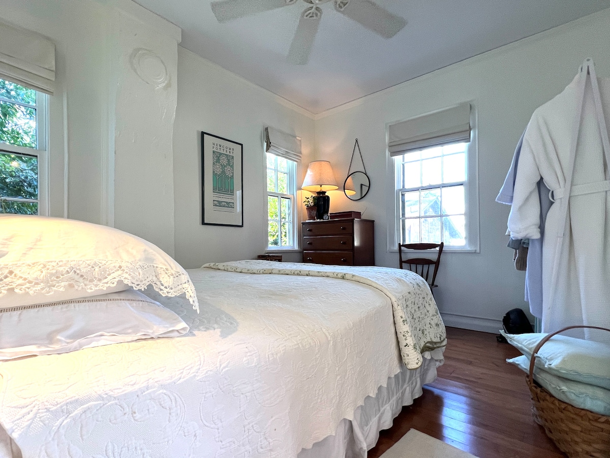 A bright bedroom features a spacious bed adorned with a decorative white quilt. Natural light enters through two windows with light curtains. A wooden dresser stands against the wall, complemented by a nearby chair and a cozy bathrobe hanging on the door.